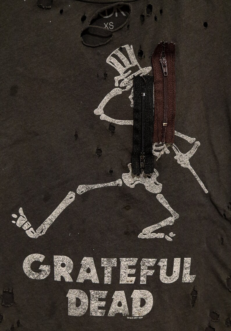 May include: A black t-shirt with a distressed look featuring a white skeleton graphic with a top hat and the text "GRATEFUL DEAD". The shirt has three zippers attached to the skeleton graphic.