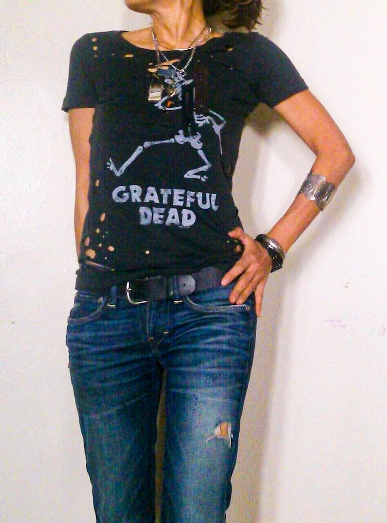 May include: A woman wearing a black t-shirt with a distressed look and a skeleton graphic. The t-shirt has the text "Grateful Dead" printed on it. She is also wearing blue jeans with a black belt.