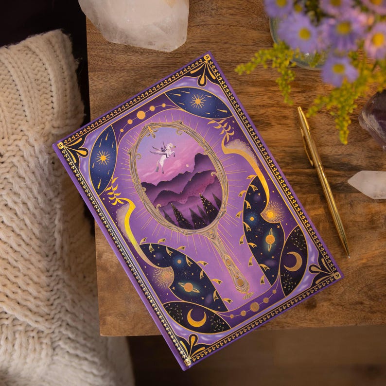 May include: Purple journal with a gold border and a design of a mirror with a unicorn flying in front of a mountain landscape. The mirror is surrounded by stars, moons, and planets.