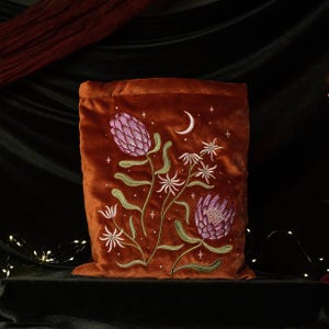 May include: A rectangular rust-colored velvet pouch featuring intricate embroidery of two large pink protea flowers, smaller white star-shaped blossoms, green stems, and a white crescent moon surrounded by tiny embroidered stars across the front.