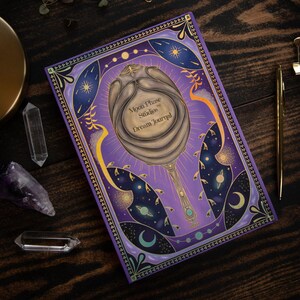 May include: Purple dream journal with a gold ornate border and a celestial illustration of a mirror with stars and planets. The text on the cover reads "Moon Phase Studios&trade; Dream Journal".