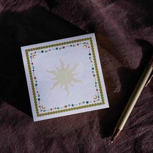 Può includere: Un blocco note quadrato con un disegno di sole al centro, circondato da un bordo decorativo di fiori, lune e stelle. Il bordo è verde con punti dorati. Una penna dorata è appoggiata su una superficie viola scuro.