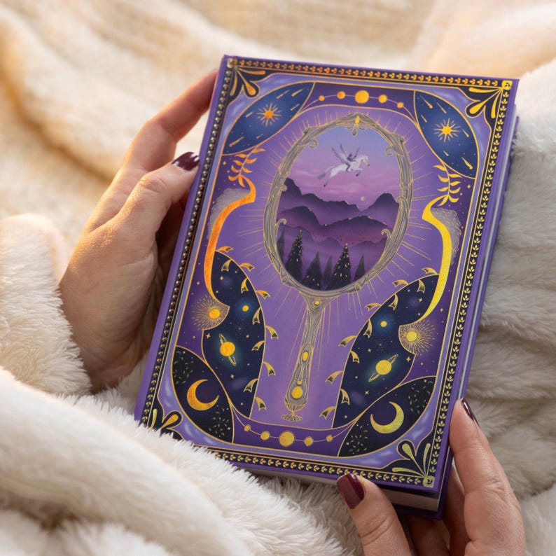 May include: Purple journal with a gold border and a design of a mirror reflecting a landscape with a unicorn flying in the sky. The mirror has a gold handle and the background is a dark purple with gold stars and moons.