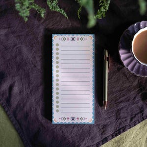 Pode incluir: Um bloco de notas retangular com uma borda azul ondulada e detalhes decorativos florais e de lua crescente. O bloco de notas tem páginas pautadas e uma série de pequenos círculos no lado esquerdo. Uma caneta e uma chávena de chá também são visíveis.
