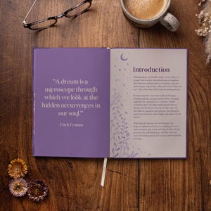 May include: Open book with purple cover and white pages. The left page has a quote about dreams by Erich Fromm. The right page has the title "Introduction" and text about the importance of dreams. The book is on a wooden table with a cup of coffee, glasses, and dried flowers.