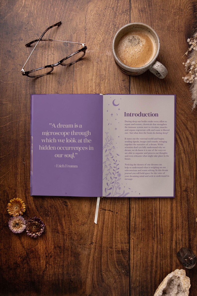 May include: Open book with purple cover and white pages. The left page has a quote about dreams by Erich Fromm. The right page has the title "Introduction" and text about the importance of dreams. The book is on a wooden table with a cup of coffee, glasses, and dried flowers.