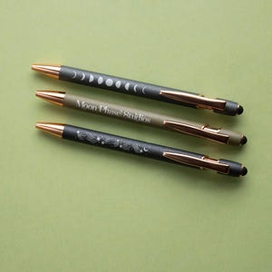 May include: Three pens with gold accents and black tips are displayed on a green surface. The top pen has a moon phase design, the middle pen reads "Moon Phase Studios", and the bottom pen has a celestial design.