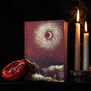 May include: A hardback book with a deep burgundy cover featuring a gold foil illustration of a swirling galaxy, a crescent moon, and a landscape. Beside the book sits a halved pomegranate and two tall, lit white taper candles with smoke.