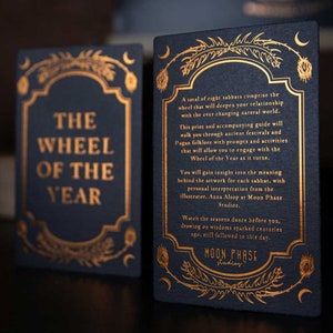 May include: A black card with gold lettering that reads "The Wheel of the Year". The card is part of a set of cards that explore the eight sabbats of the Wheel of the Year. The card is illustrated with a stylized floral design.