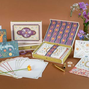 May include: A stationery set with a celestial design, including a box, envelopes, and cards. The box is open, revealing the stationery inside, and features a purple and gold colour scheme. A gold pen and flower are also present.