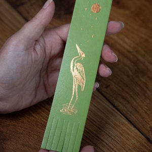 May include: Green leather bookmark with a gold foil design of a heron standing in water with a moon and stars in the background.