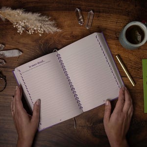 May include: An open spiral bound journal with purple accents and lined pages. The journal is open to a page that says "In more detail..." and has a gold pen and a green bookmark with a bird design next to it.