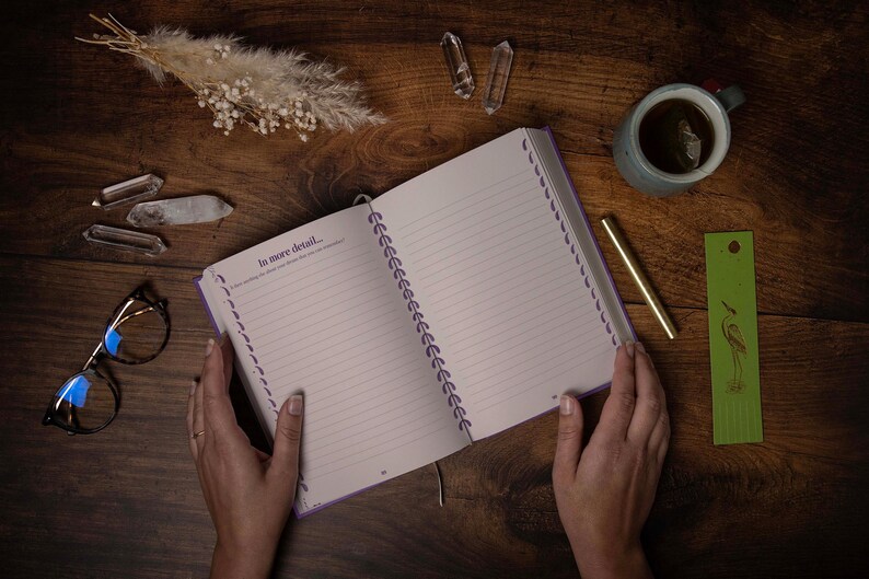 May include: An open spiral bound journal with purple accents and lined pages. The journal is open to a page that says "In more detail..." and has a gold pen and a green bookmark with a bird design next to it.