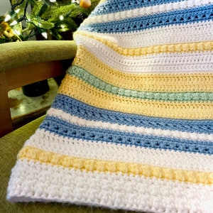 May include: A striped crocheted baby blanket in white, yellow, light blue, and green. The blanket is folded and draped over a green chair.