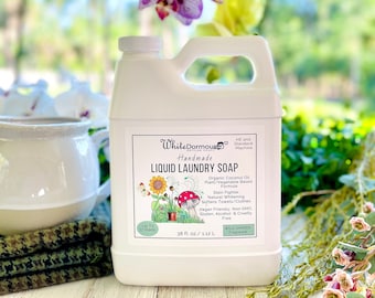 May include: White Dormouse Handmade Liquid Laundry Soap in a white plastic bottle with a green and yellow label. The label features a cartoon illustration of a sunflower, mushrooms, and a ladybug. The bottle is sitting on a wooden surface with a white pitcher and green and brown fabric in the background. The label text reads: "HE and Standard Machine", "Handmade", "Liquid Laundry Soap", "Organic Coconut Oil Plant/Vegetable Based Formula", "Stain Fighter", "Natural Whitening", "Softens Towels/Clothes", "Vegan Friendly, Non GMO, Gluten, Alcohol & Cruelty Free", "Up to 17 Loads", "38 fl. oz./1.12 L", "Wild Garden Fragrance".