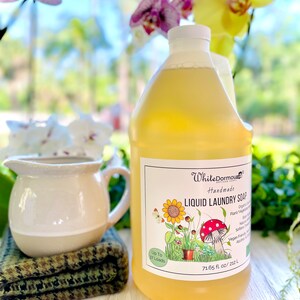 May include: A large, clear plastic bottle of yellow liquid laundry soap with a white label. The label has a cartoon illustration of a sunflower, a mushroom, and a potted plant. The label also says "White Dormouse Handmade Liquid Laundry Soap" and "Up to 37 Loads".