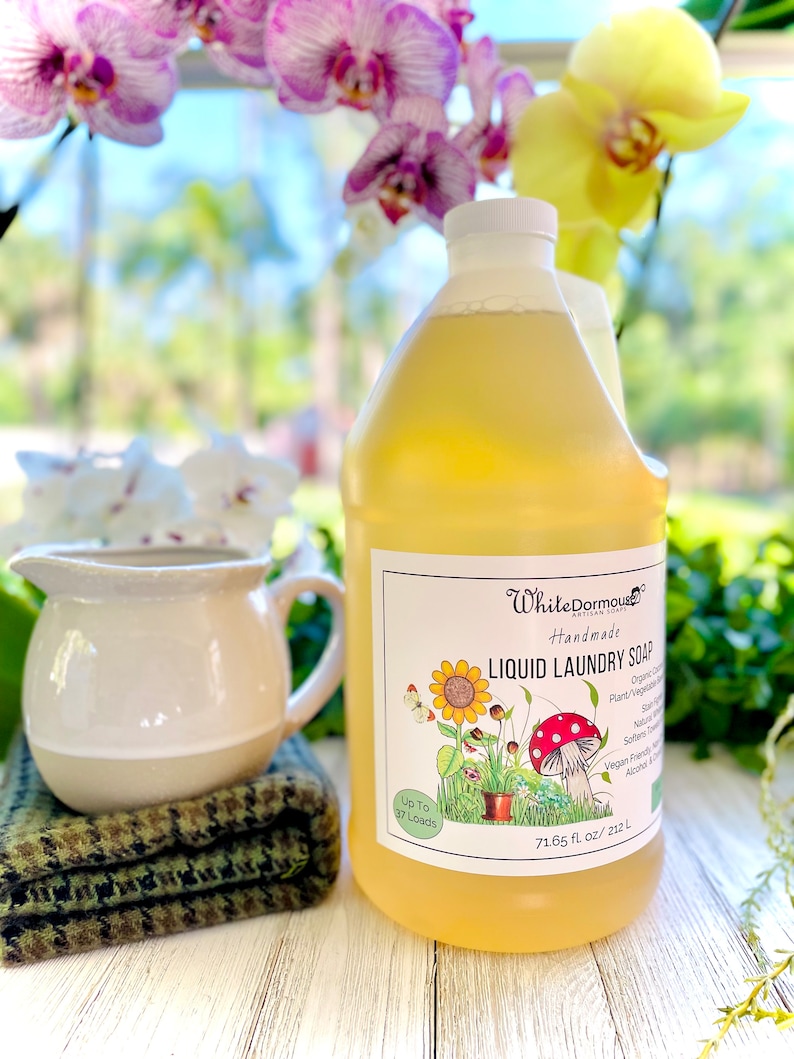 May include: A large, clear plastic bottle of yellow liquid laundry soap with a white label. The label has a cartoon illustration of a sunflower, a mushroom, and a potted plant. The label also says "White Dormouse Handmade Liquid Laundry Soap" and "Up to 37 Loads".