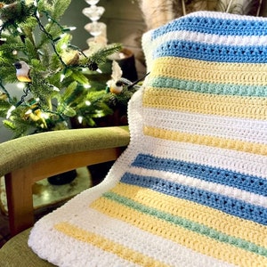 May include: A white, yellow, blue and green striped crocheted baby blanket. The blanket is folded and draped over a green chair.