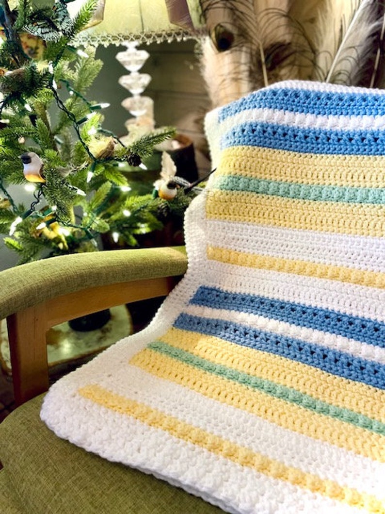 May include: A white, yellow, blue and green striped crocheted baby blanket. The blanket is folded and draped over a green chair.