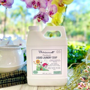 May include: White Dormouse Handmade Liquid Laundry Soap in a white plastic jug with a green label. The label features a cartoon illustration of a sunflower, mushrooms, and a garden. The label also includes the text "Up to 17 loads", "38 fl oz / 1.12 L", "Wild Garden Fragrance", and a list of product features.