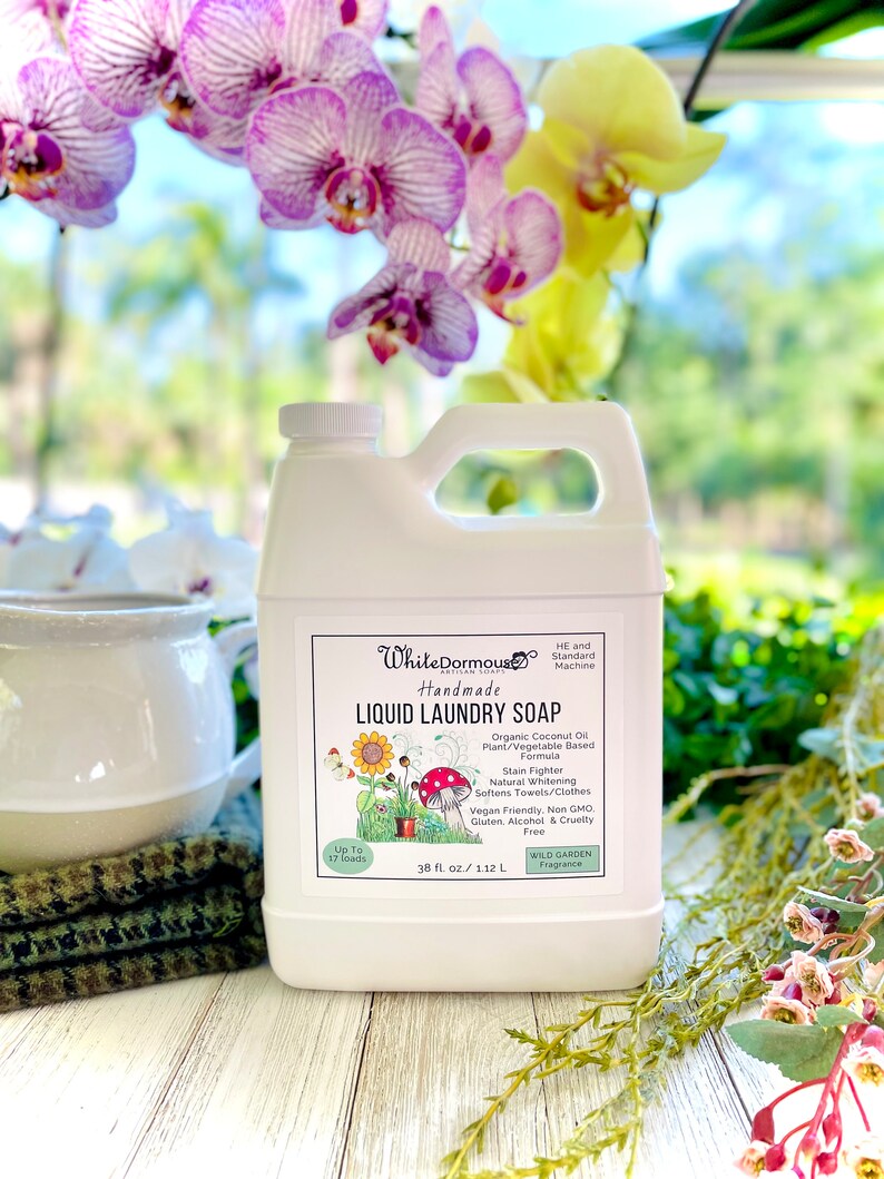May include: White Dormouse Handmade Liquid Laundry Soap in a white plastic jug with a green label. The label features a cartoon illustration of a sunflower, mushrooms, and a garden. The label also includes the text "Up to 17 loads", "38 fl oz / 1.12 L", "Wild Garden Fragrance", and a list of product features.