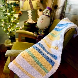 May include: A crocheted baby blanket with stripes of white, yellow, blue, and green. The blanket is draped over a rocking chair.
