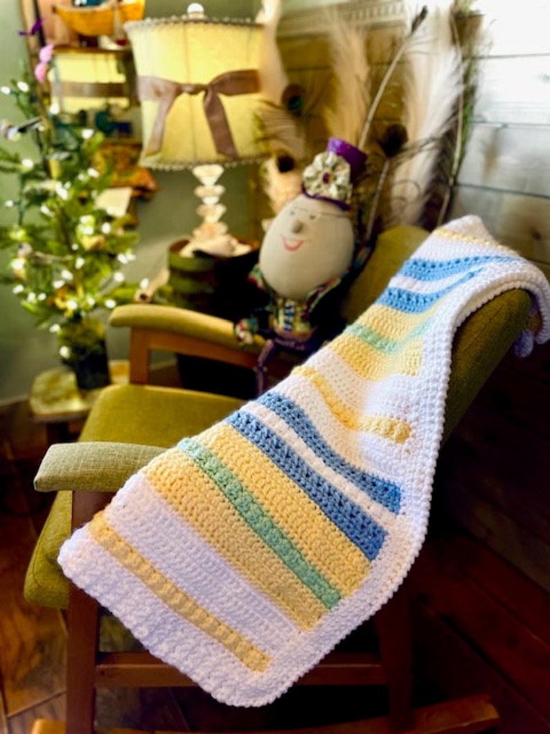 May include: A crocheted baby blanket with stripes of white, yellow, blue, and green. The blanket is draped over a rocking chair.