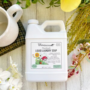May include: White Dormous Handmade Liquid Laundry Soap in a white plastic jug with a green label. The label features a sunflower and mushroom illustration. The soap is made with organic coconut oil and is plant and vegetable based. It is a stain fighter, naturally whitens, softens towels and clothes, is vegan friendly, non-GMO, gluten, alcohol and cruelty free. It has a Wild Garden fragrance. The jug contains 38 fl oz or 1.12 L.