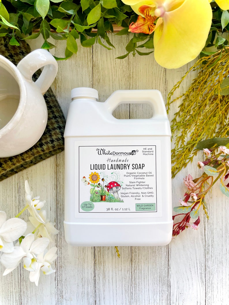 May include: White Dormous Handmade Liquid Laundry Soap in a white plastic jug with a green label. The label features a sunflower and mushroom illustration. The soap is made with organic coconut oil and is plant and vegetable based. It is a stain fighter, naturally whitens, softens towels and clothes, is vegan friendly, non-GMO, gluten, alcohol and cruelty free. It has a Wild Garden fragrance. The jug contains 38 fl oz or 1.12 L.