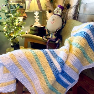 May include: A striped crocheted baby blanket in white, yellow, blue, and green. The blanket is draped over a rocking chair with a doll wearing a crown sitting in the chair.