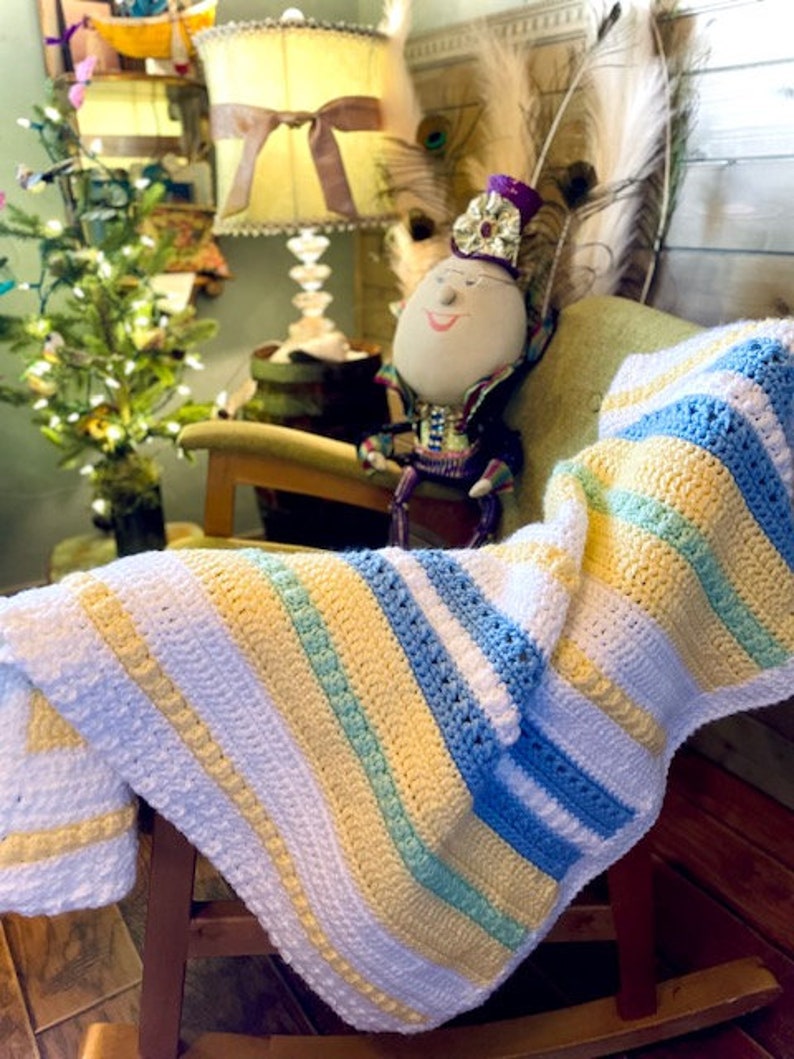 May include: A striped crocheted baby blanket in white, yellow, blue, and green. The blanket is draped over a rocking chair with a doll wearing a crown sitting in the chair.