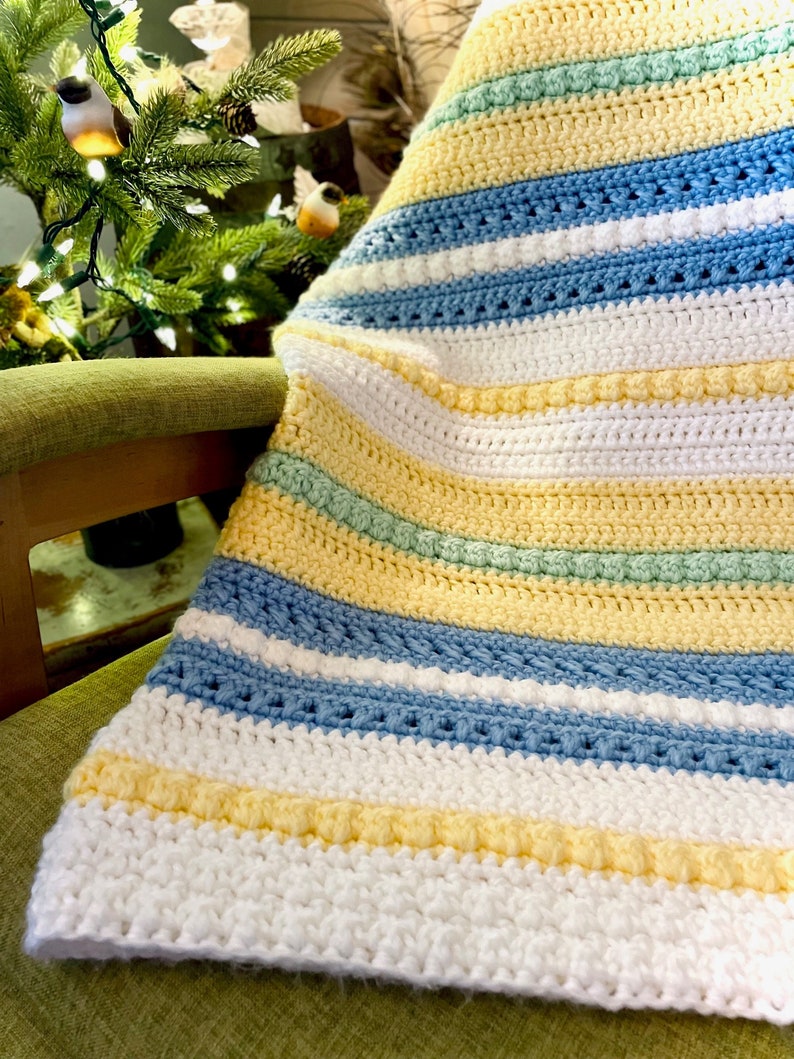 May include: A striped crocheted baby blanket in white, yellow, light blue, and green. The blanket is folded and draped over a green chair.