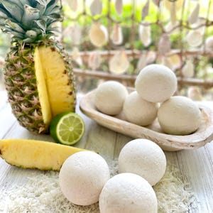 May include: A halved pineapple with a slice, a lime wedge, and several white, round bath bombs are arranged on a white wooden surface. Shredded coconut is scattered around the bath bombs. A wooden bowl holds more bath bombs.