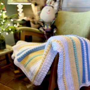 May include: A white crocheted baby blanket with blue, yellow and green stripes. The blanket is draped over a wooden rocking chair.