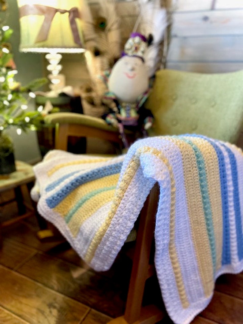 May include: A white crocheted baby blanket with blue, yellow and green stripes. The blanket is draped over a wooden rocking chair.