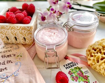 May include: Two glass jars filled with pink raspberry body scrub, a basket of fresh raspberries, a pink and white floral arrangement, and a sign that says "Farmer's Market, Open Daily, You Pick Raspberries".