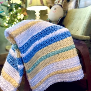 May include: A striped crocheted baby blanket in shades of white, yellow, blue, and green. The blanket is draped over a rocking chair.