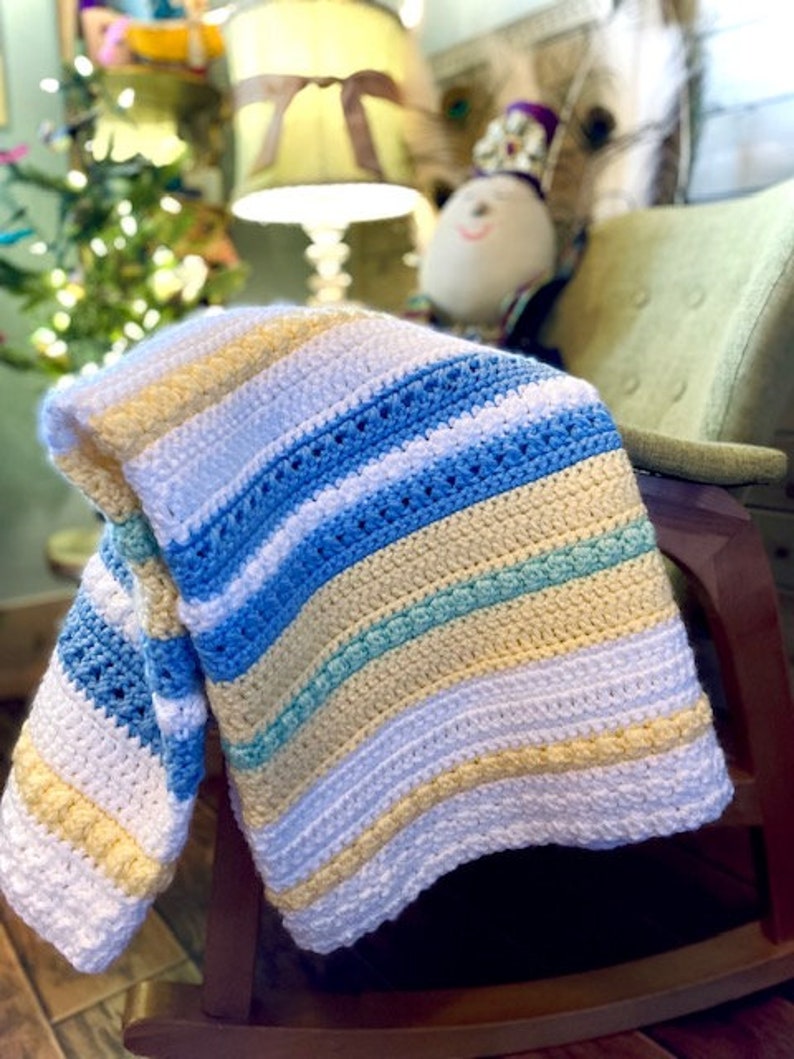 May include: A striped crocheted baby blanket in shades of white, yellow, blue, and green. The blanket is draped over a rocking chair.