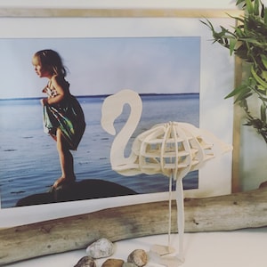 May include: A white wooden flamingo sculpture with a lattice design. The sculpture is standing on a wooden surface with a few small rocks.