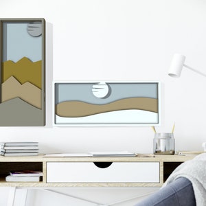 May include: Two framed paper cut-out art pieces depicting a landscape with mountains and a moon. The art pieces are hanging on a white wall above a wooden desk with a drawer.