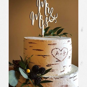 May include: Two-tiered wedding cake decorated with birch bark frosting and greenery. The top tier has a carved heart with the initials "A+R". The cake topper is a wooden sign that says "Mr & Mrs".