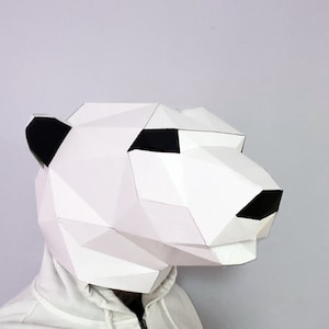 May include: A white papercraft mask of a polar bear head. The mask is made of folded paper and has black eyes and a black nose.