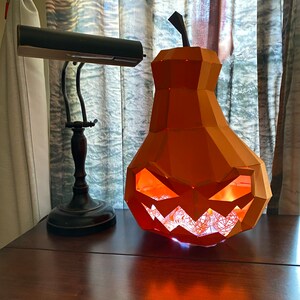 May include: A papercraft pumpkin shaped like a pear with a carved jack-o'-lantern face. The pumpkin is lit from inside with white fairy lights, casting a warm glow.