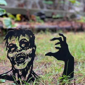 May include: Black metal silhouette of a zombie head and a hand reaching out, both placed on a grassy surface.