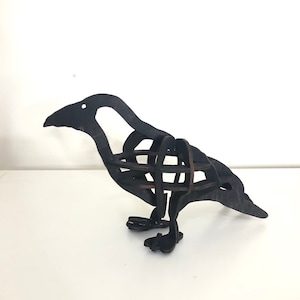 Könnte beinhalten: Eine schwarze Metallsculptur eines Rabens mit einem Drahtkäfig-Design. Der Rabe steht auf einer weißen Oberfläche.