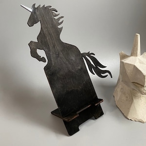 Puede incluir: Un soporte de teléfono de madera negra con forma de silueta de unicornio. El soporte tiene una pequeña plataforma elevada para sostener un teléfono.