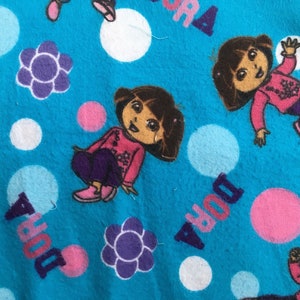May include: Blue fabric with a repeating pattern of Dora the Explorer, pink and white polka dots, and purple flowers. The text "Dora" is repeated throughout the fabric.