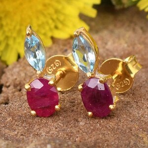 Puede incluir: Un par de pendientes de botón dorados con una piedra preciosa azul claro en talla marquesa sobre una piedra roja oscura en talla redonda. Cada pendiente tiene el sello 925 en la parte posterior, indicando plata de ley con acabado dorado.