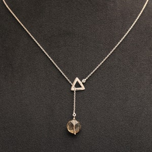 Op de afbeelding: Een zilveren ketting met een driehoekige hanger en een bruine edelsteen die eronder hangt.