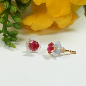 May include: A pair of stud earrings featuring round, faceted, deep pink stones set in gold-toned metal prongs. Each earring includes a clear, circular plastic backing to secure the post, providing a simple and functional design for ear jewellery.